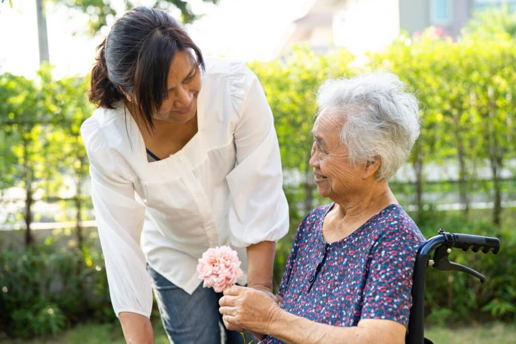 Caregiver and elderly woman holding flower, smiling and happy in the sunny garden.